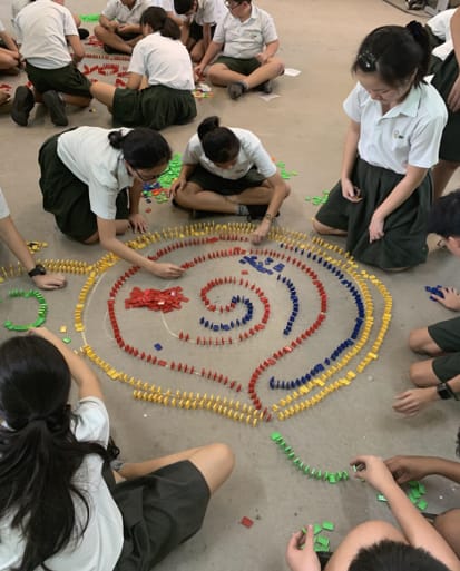 Students in school uniforms sit on a floor arranging colorful dominoes in a large spiral pattern.